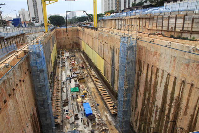 High-contrast duotone photograph in deep charcoal and pale teal showing an aerial view of a rectangular construction excavation with braced walls, workers, and equipment set against distant city buildings.
